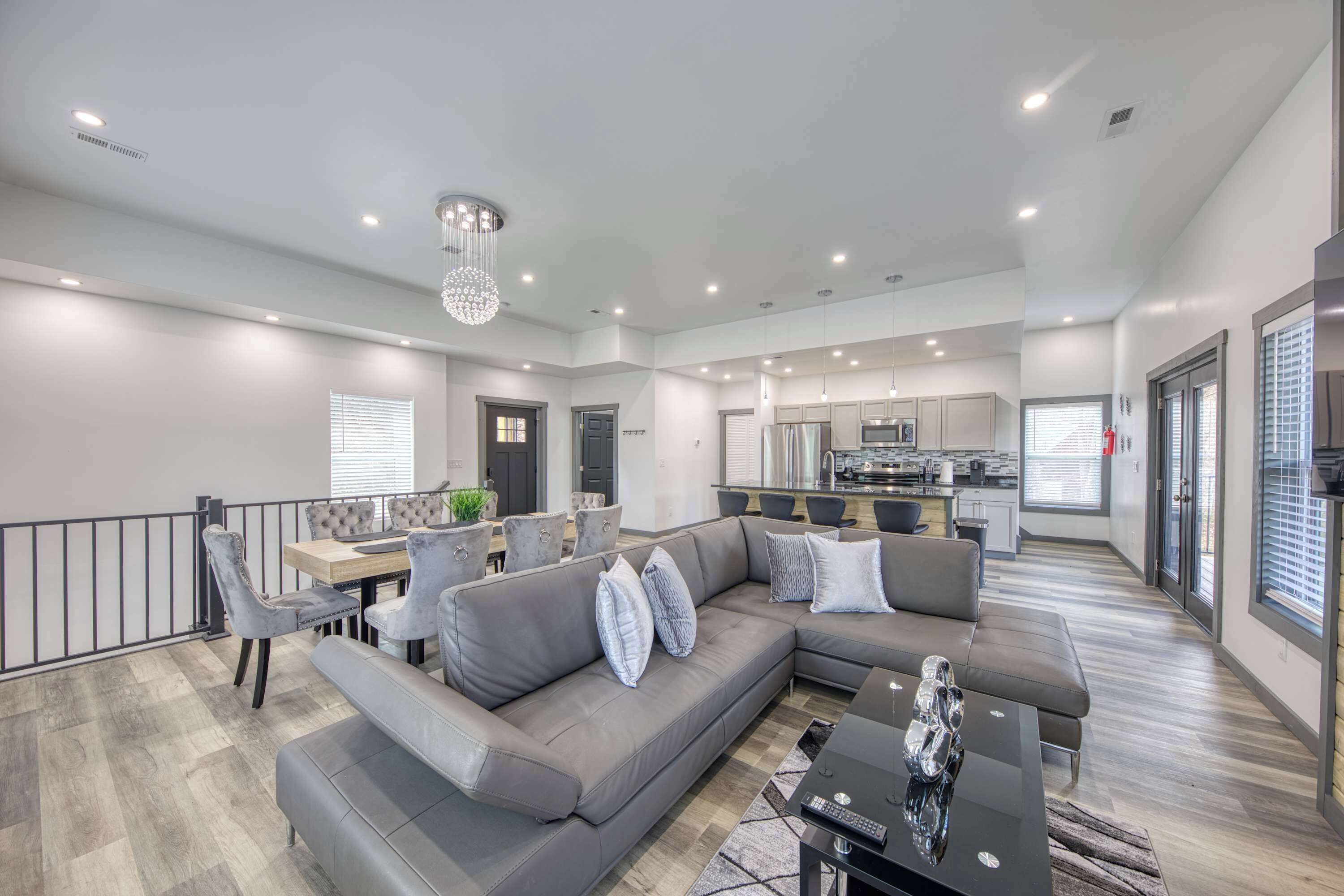 A spacious living area with a grey sectional sofa, a dining table with chairs, a kitchen in the background, and a balcony with a railing.