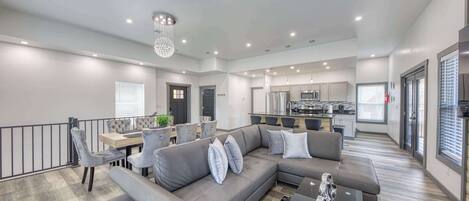 A spacious living area with a grey sectional sofa, a dining table with chairs, a kitchen in the background, and a balcony with a railing.
