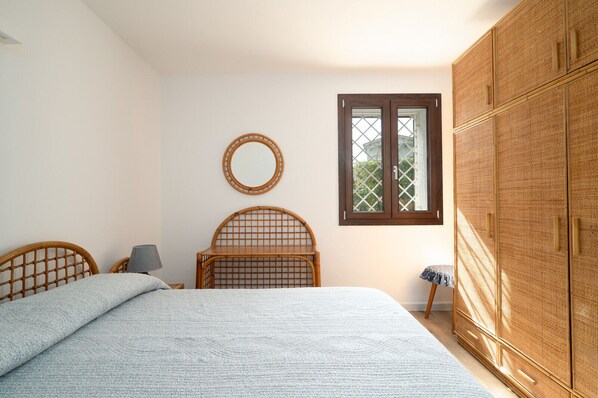 A bedroom with a bed, a wooden wardrobe, a round mirror, a window, and a small table.