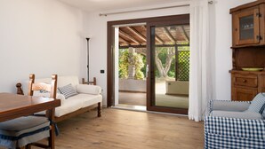 A room with a wooden table, a sofa, a cabinet, and a sliding glass door leading to an outdoor area with a wooden pergola and greenery.