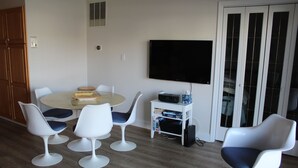 A dining area with a round table, six white chairs, and a flat-screen TV mounted on the wall.