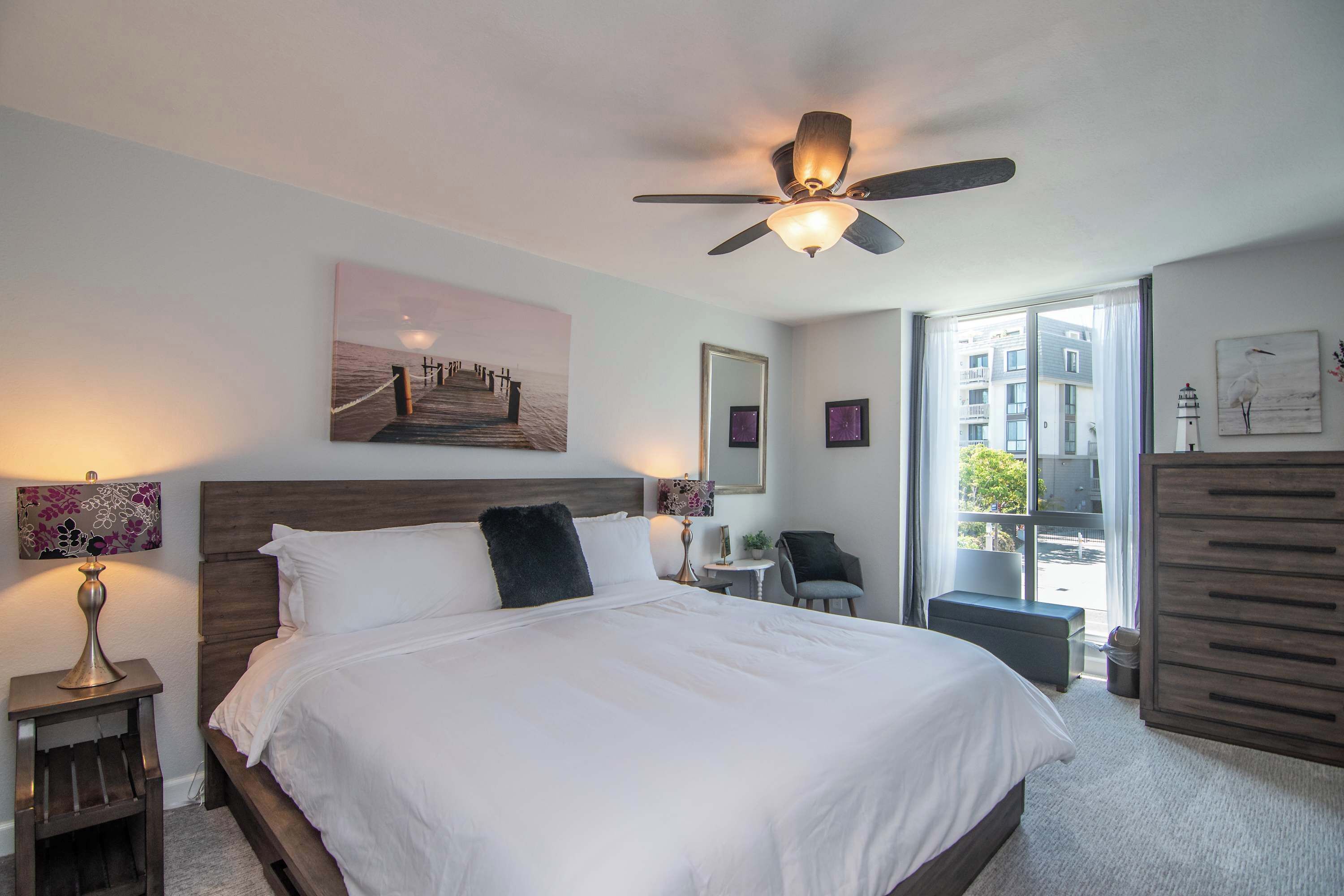 Habitación de hotel con una cama grande, ventilador de techo, escritorio con silla, ventana con vistas a edificios y cómoda.