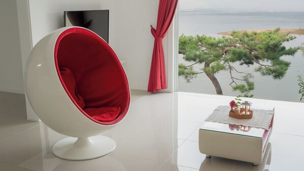 Una habitación moderna con una silla blanca, una mesita con un objeto decorativo, un televisor y vistas a un cuerpo de agua y árboles.
