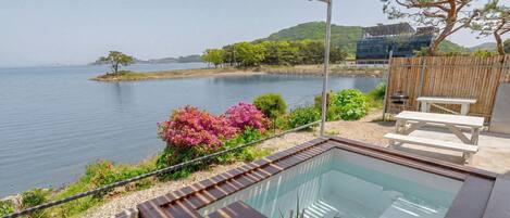 A pool with a wooden deck, a covered seating area, and a view of the water and hills.