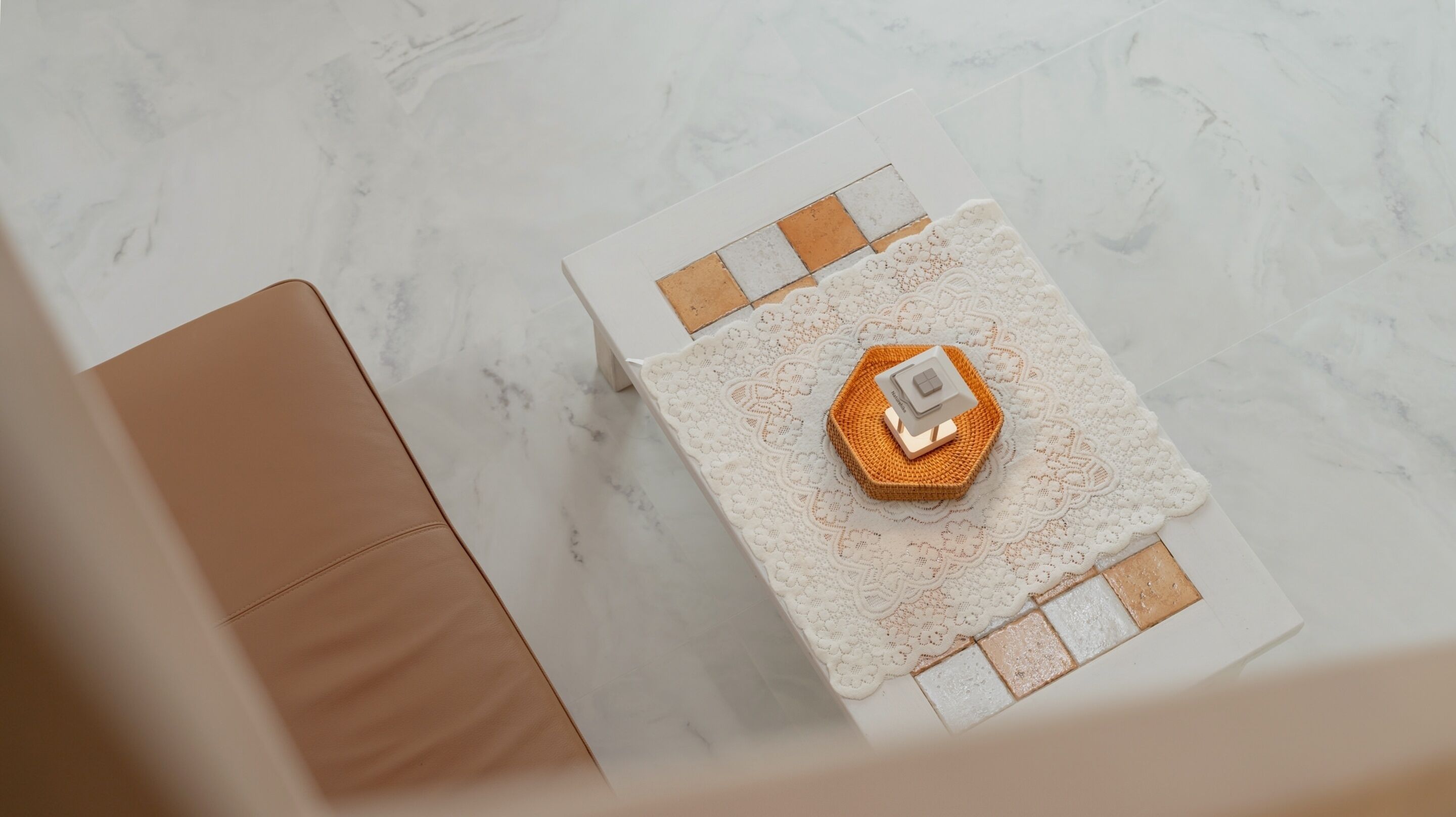 Vista cenital de una habitación con piso de mármol blanco, una mesa blanca con mantel tejido y una silla de cuero marrón.