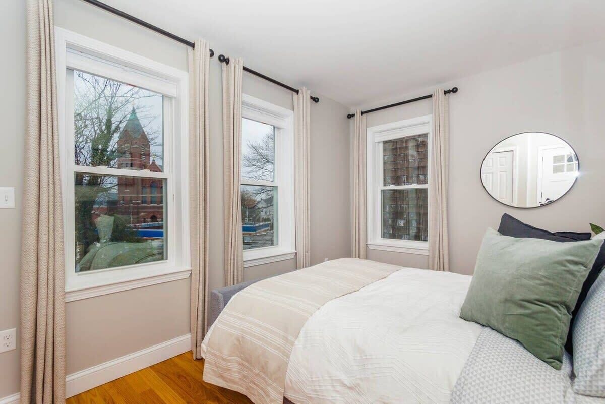 Une chambre à coucher avec un lit, deux fenêtres, un miroir et une vue sur un immeuble en briques à l’extérieur.