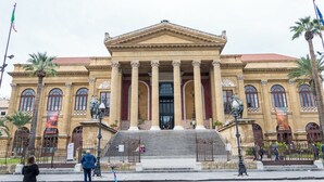 A classical building with a central entrance, flanked by columns, and palm trees in the foreground.