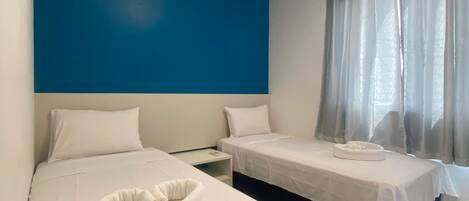 A hotel room with two beds, a blue accent wall, an air conditioning unit, a bedside table, and a window with sheer curtains.