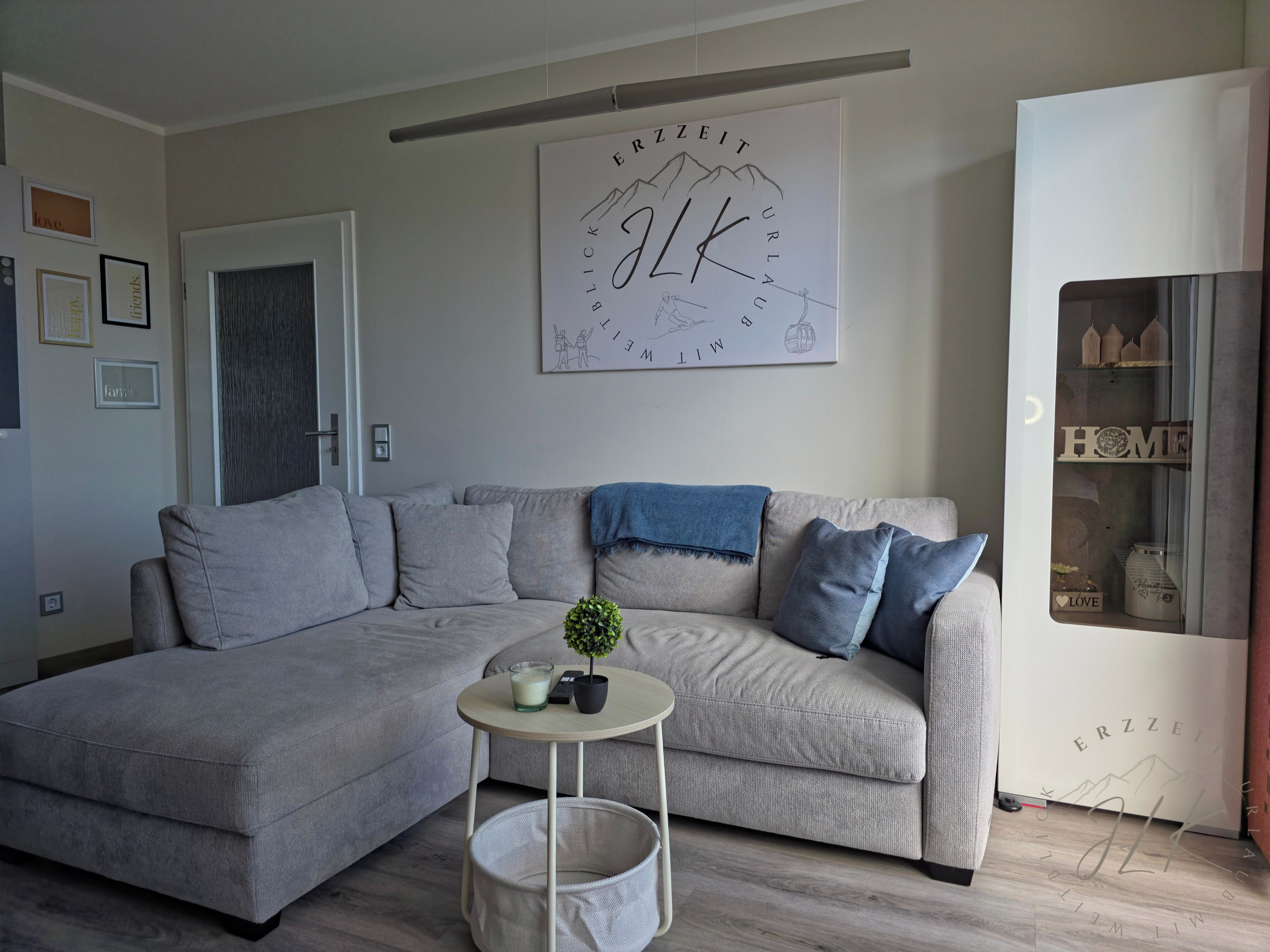 Ein modernes Wohnzimmer mit einer grauen Eckcouch, einem kleinen runden Tisch mit Kerze und Pflanze sowie einem Wandbild mit Berglandschaft und dem Wort "ALTI".