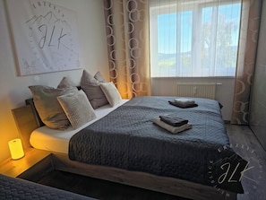 A bedroom with a bed, pillows, a bedside table with a lamp, a window with curtains, and a wall with a poster.