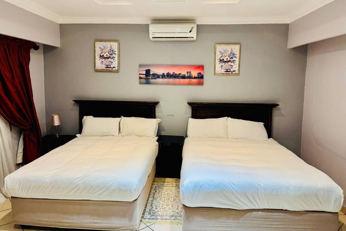 A hotel room with two single beds, a cityscape painting on the wall, and an air conditioning unit.