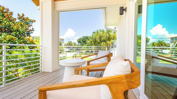 A wooden deck with a white table and two cushioned chairs, overlooking a lush garden and a clear blue sky. - Spacious Pensacola Beach Retreat with Private Pool & Game Room | Sleeps 15 (Pensacola Beach)