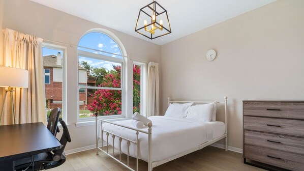 A bedroom with a bed, a desk, a chair, a dresser, and a window with a view of pink flowers and a brick building.