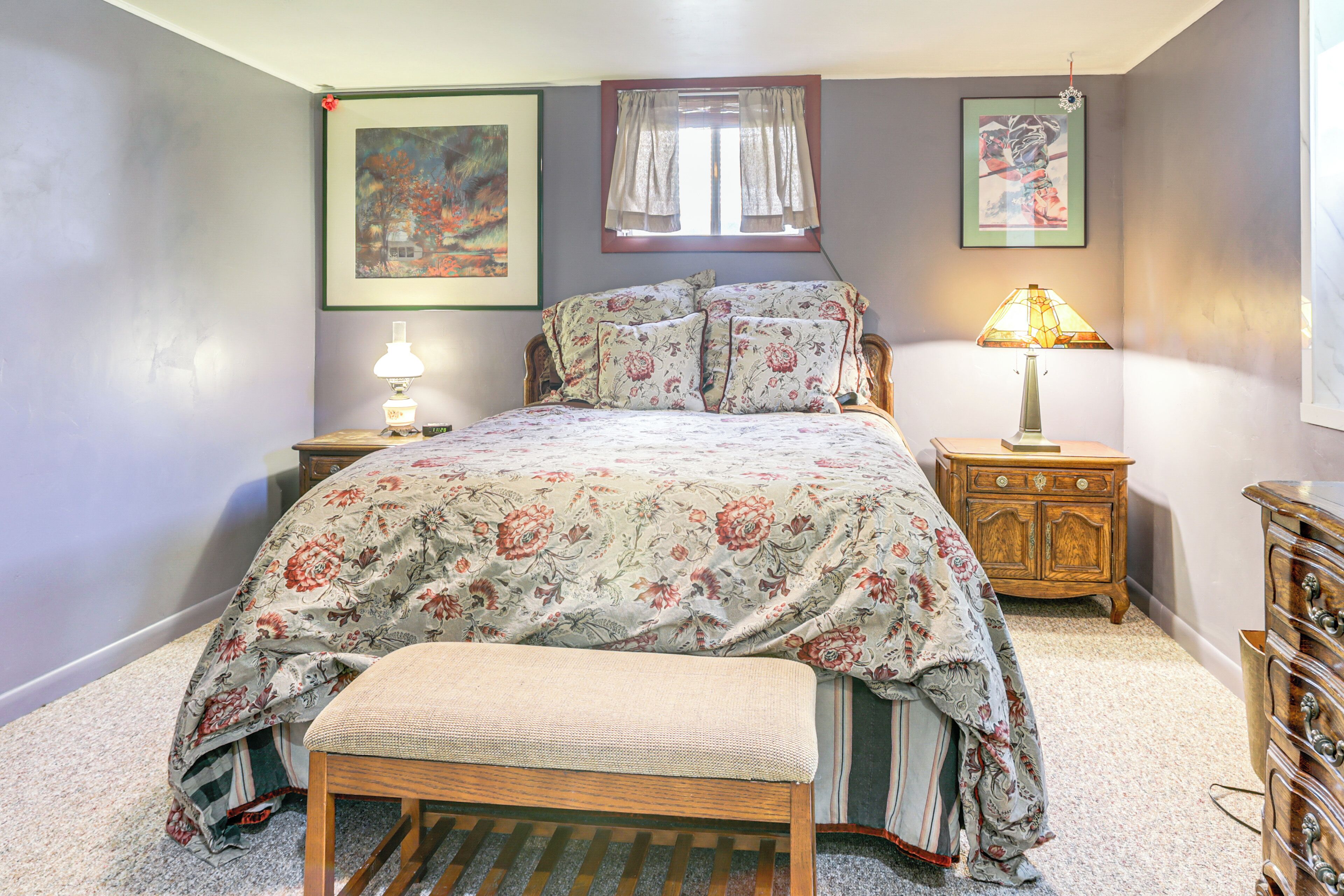 A bedroom with a floral bedspread, a wooden bench, a nightstand with a lamp, and a dresser.