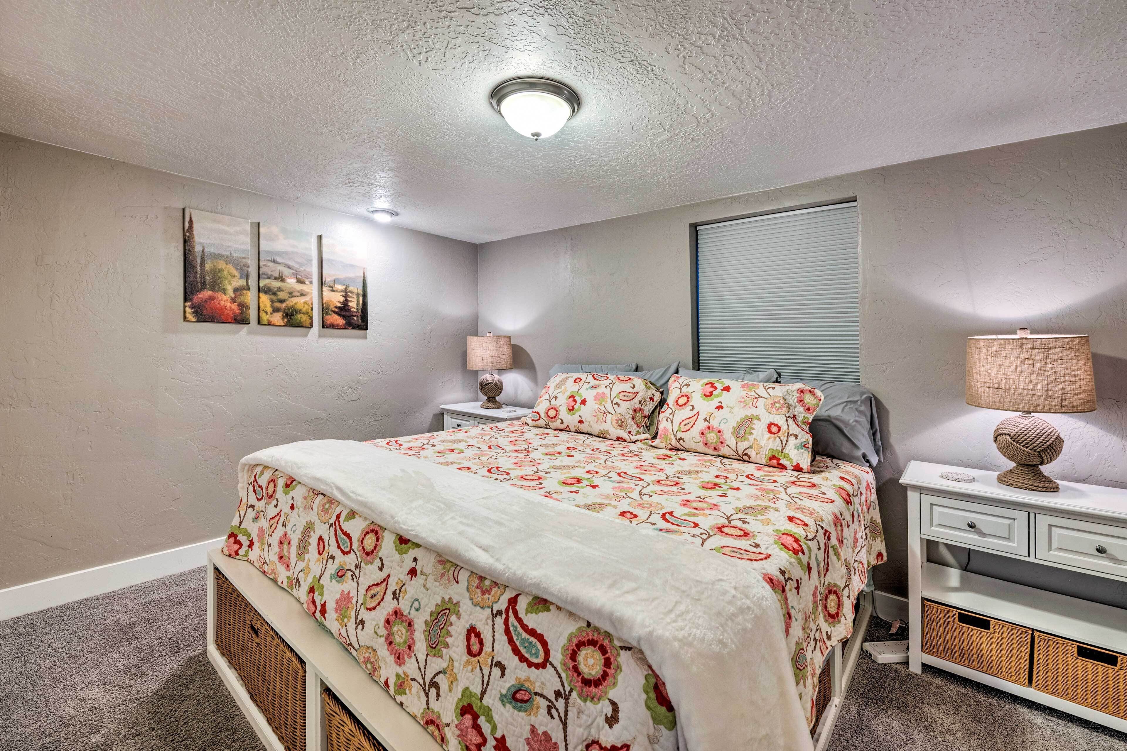 A bedroom with a floral bedspread, a bedside lamp, a nightstand, and a window with blinds.