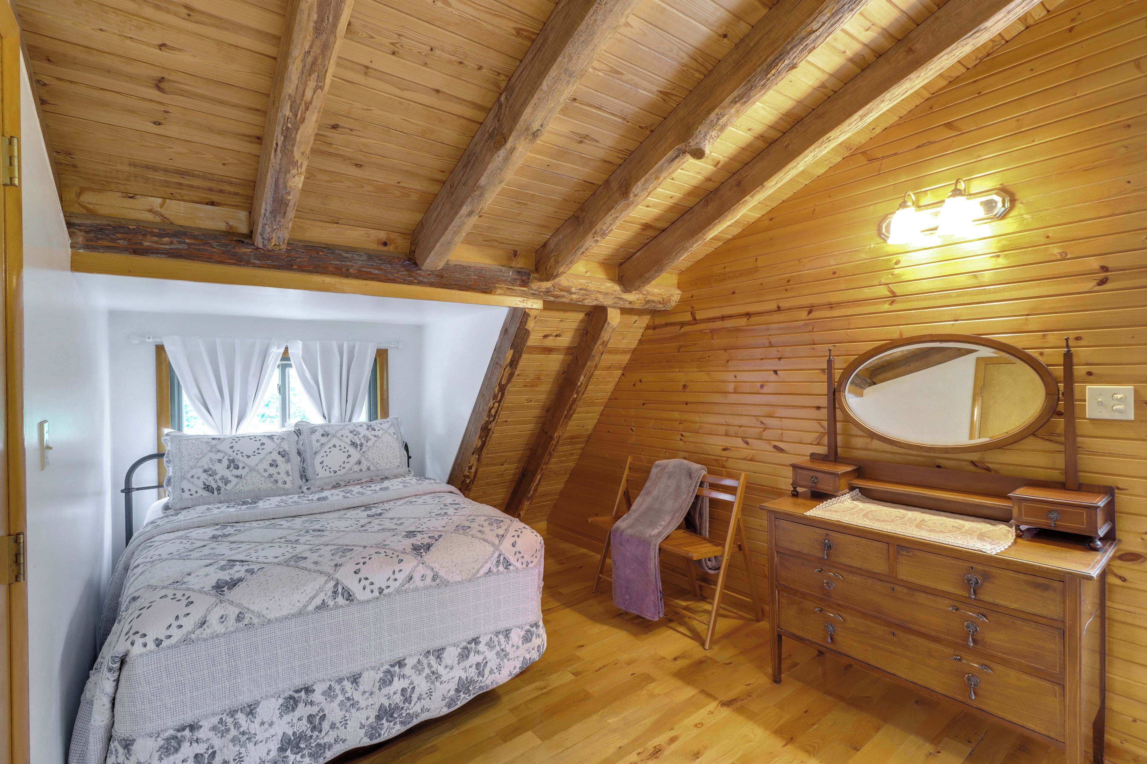 Ein gemütliches Schlafzimmer mit Holzbalkendecke, einem Bett mit floralem Überwurf, einem Schminktisch mit Spiegel und einem Fenster mit Vorhängen.
