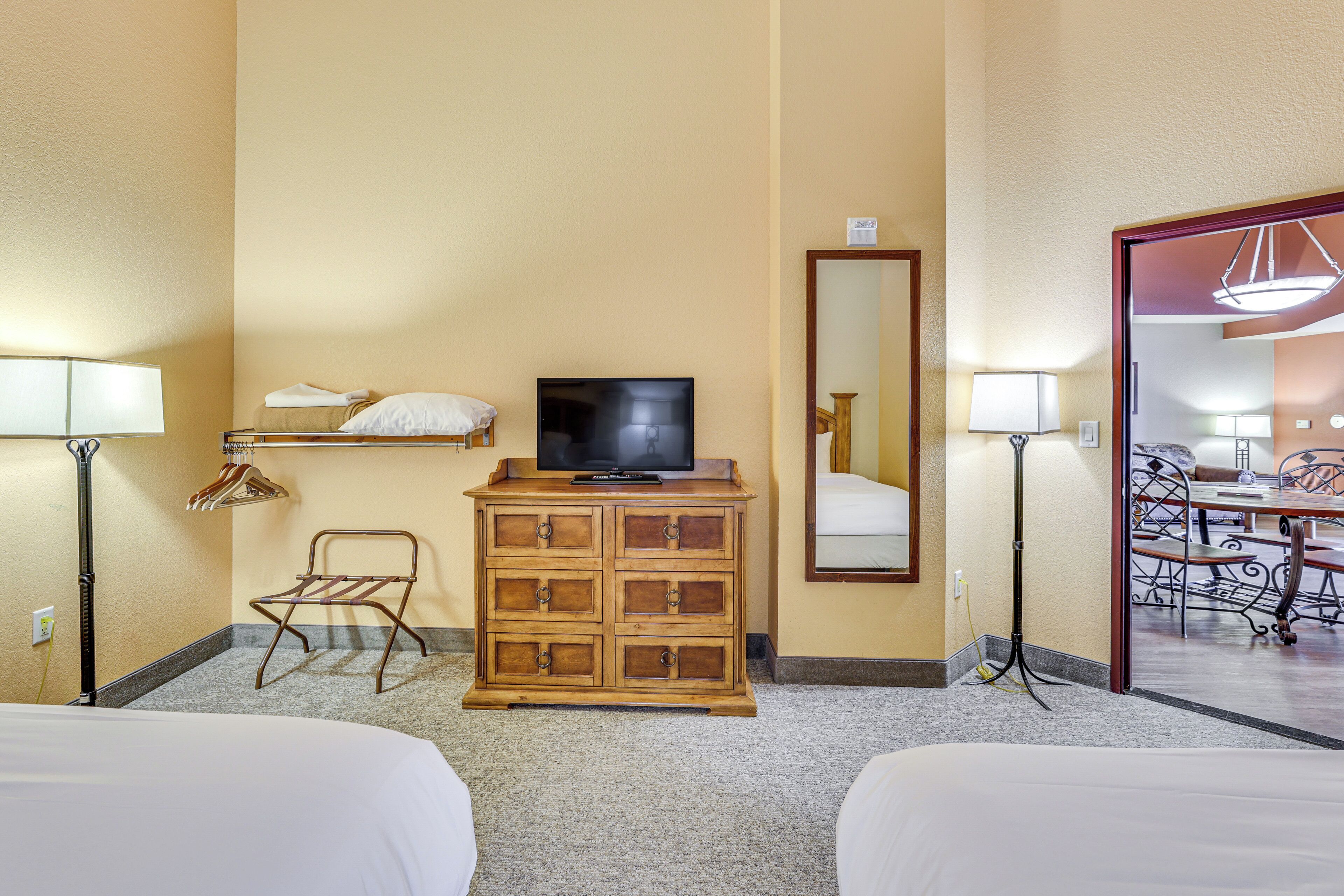 A hotel room with two beds, a wooden dresser with a television, a mirror, and a dining area visible through an open door.