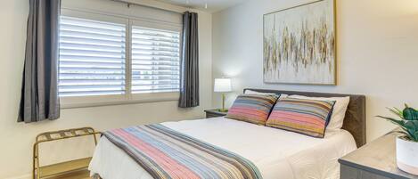 A bedroom with a bed, a ceiling fan, a window with curtains, a bedside table with a lamp, and a painting on the wall.
