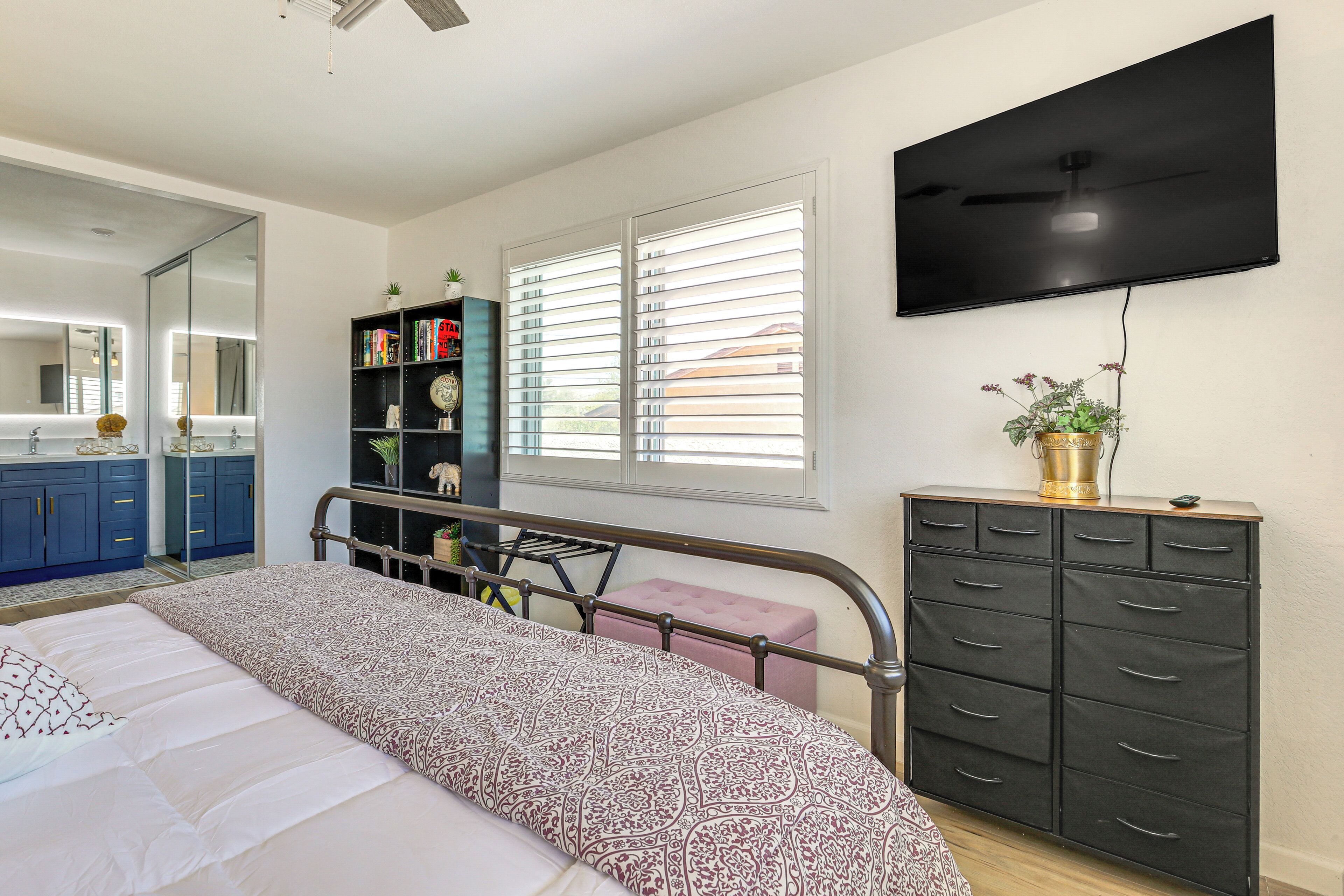 Un dormitorio con cama, armario, televisor, ventana con persianas y baño con espejo y lavamanos.