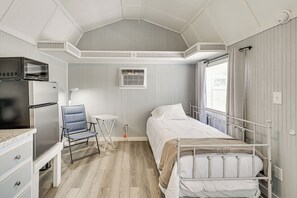 Une petite chambre comprenant un lit, un petit bureau, une chaise, un réfrigérateur et une fenêtre avec des rideaux.