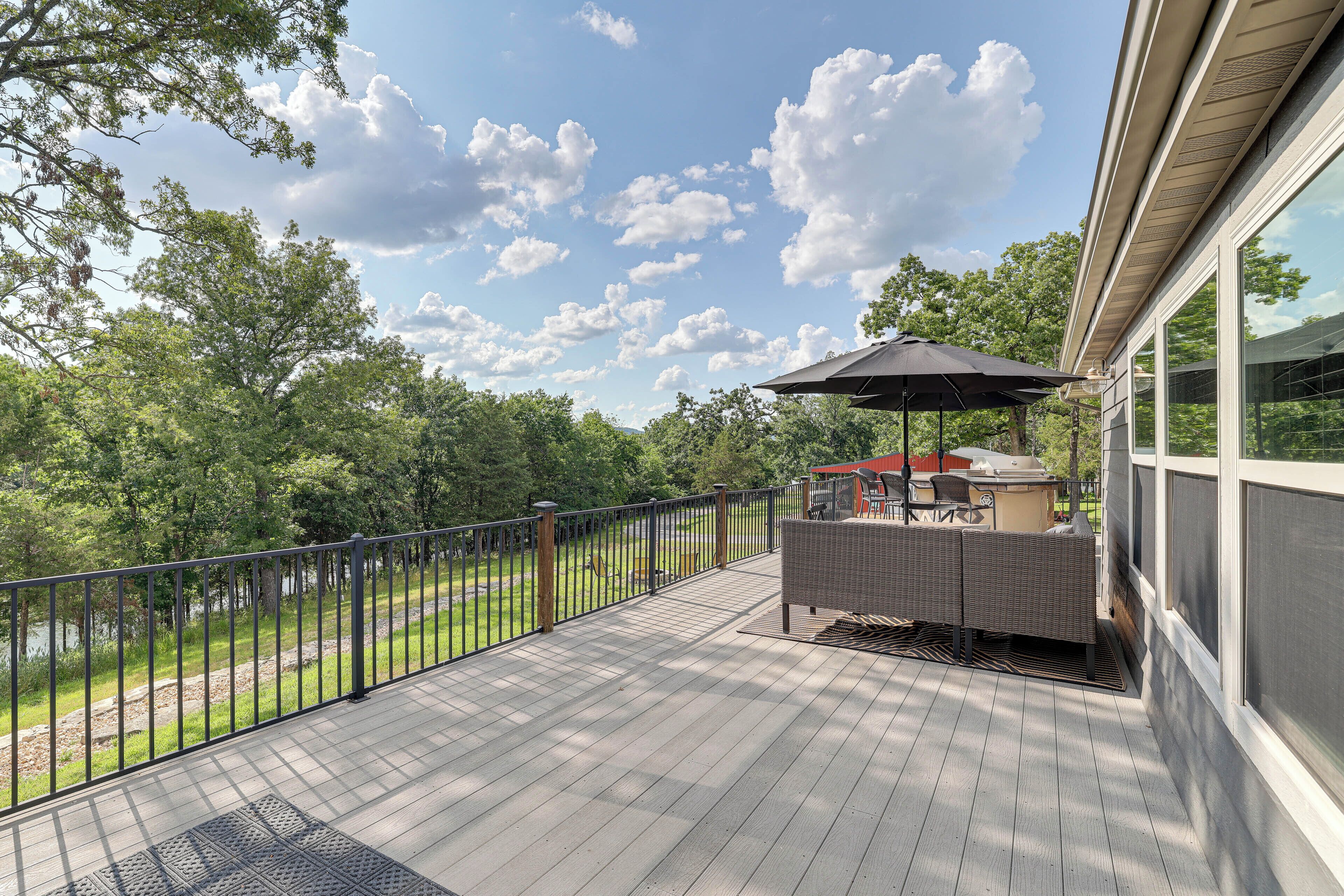 Una terraza de madera con barandilla de metal, mobiliario exterior y vista a zona boscosa.