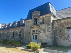 A historic stone building with a central entrance, multiple windows, and a small garden area with a bench.