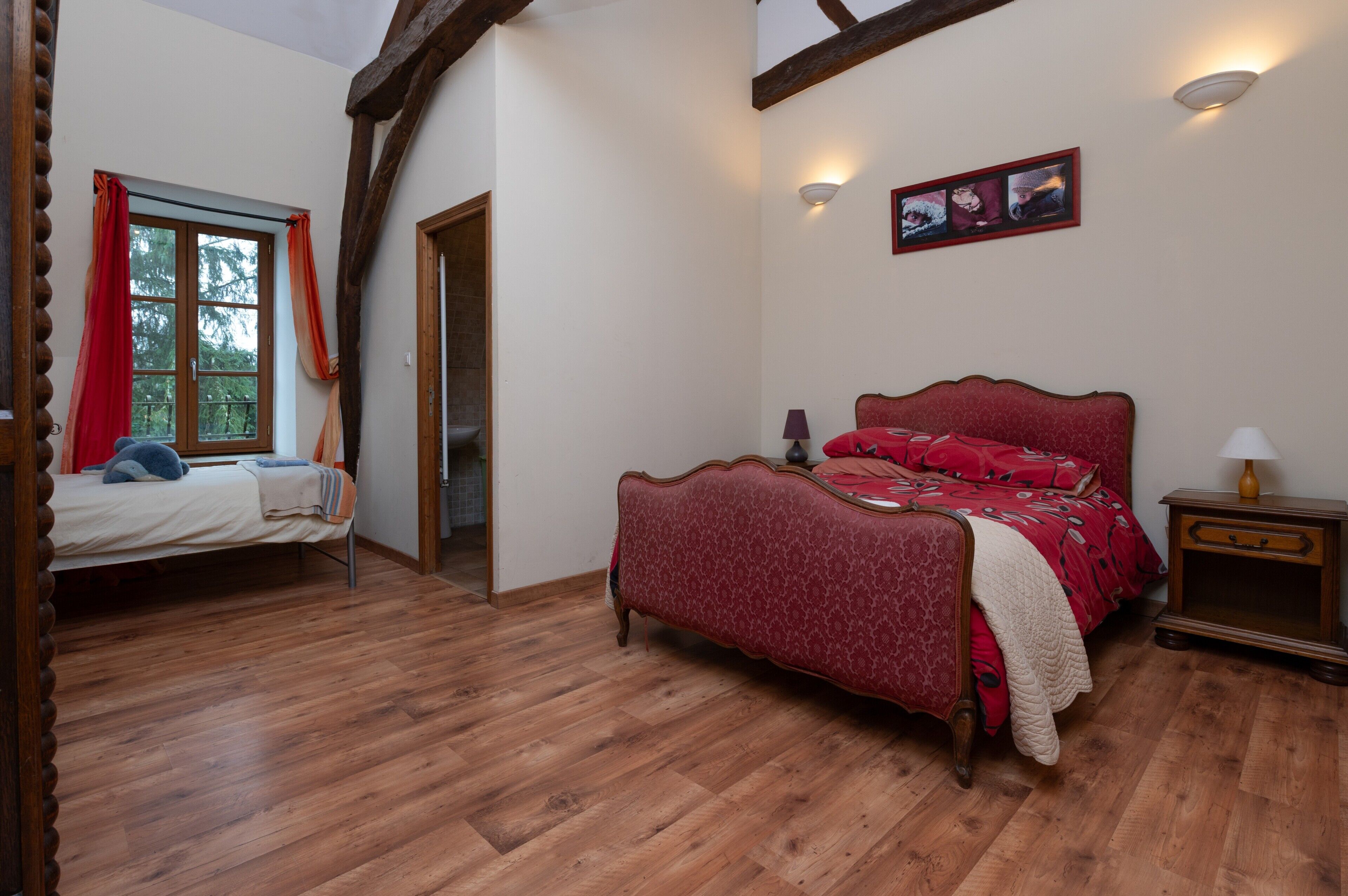 A bedroom with a wooden floor, a bed with red bedding, a nightstand, and a wall-mounted picture.