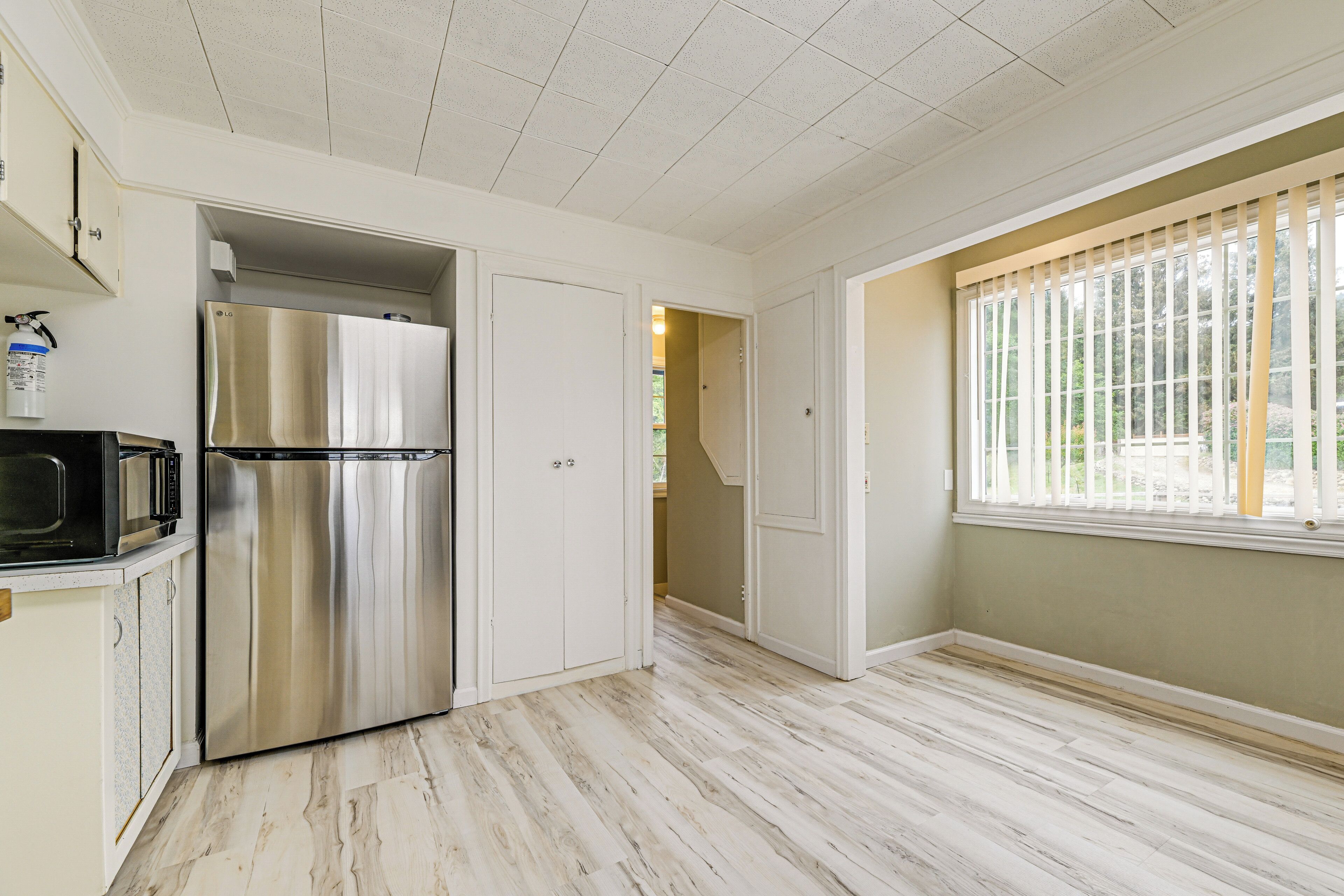 Una cocina con un refrigerador de acero inoxidable, un microondas y un lavamanos. Hay una ventana con persianas y una puerta que da a otra habitación.