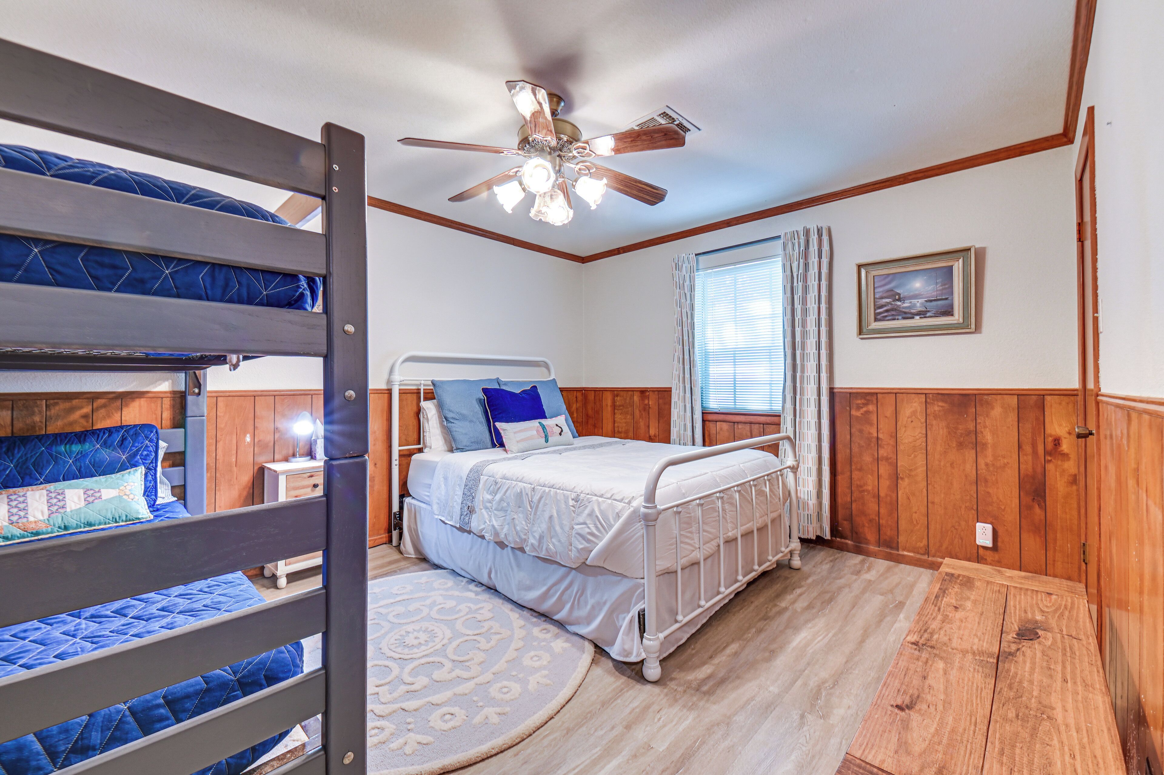 A bedroom with bunk beds, a single bed, a ceiling fan, a window with curtains, and a wooden floor.