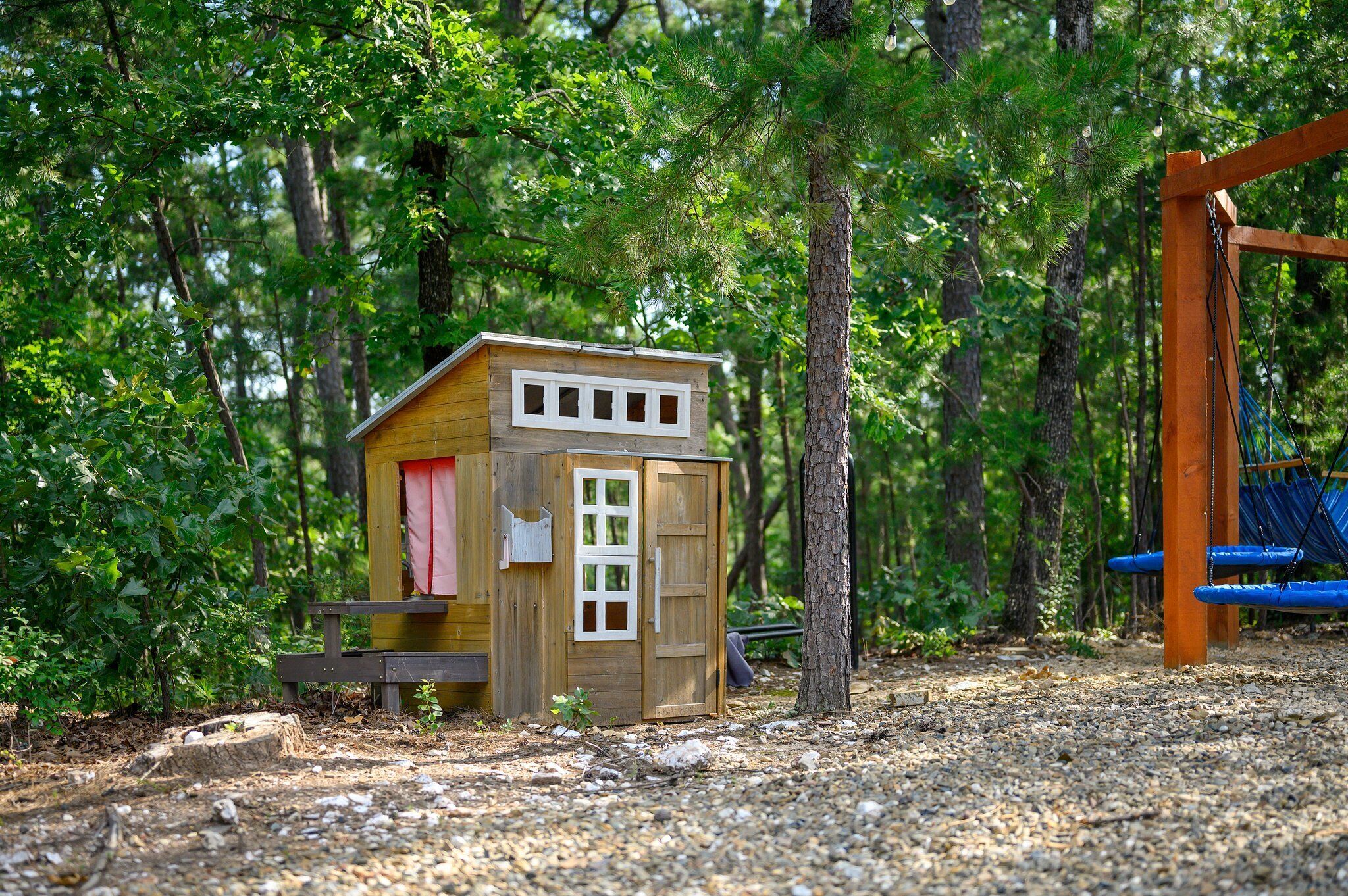 A wooden playhouse with a red door and windows, surrounded by trees and a swing set.