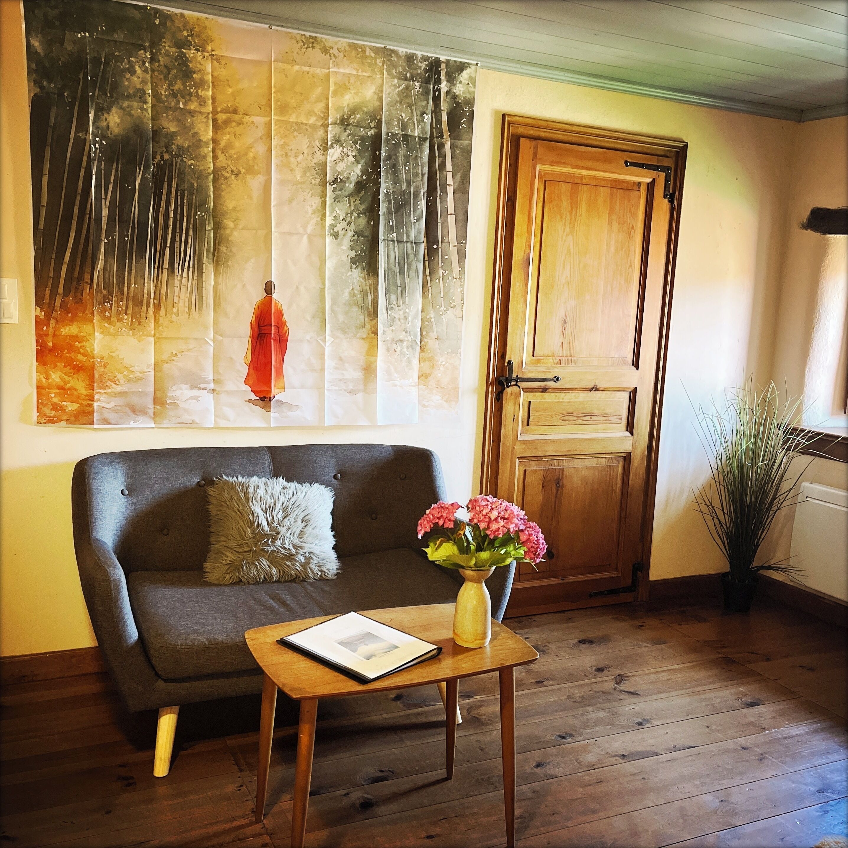 A room with a dark grey sofa, a small wooden table with a vase of pink flowers, and a large painting on the wall depicting a serene forest scene.
