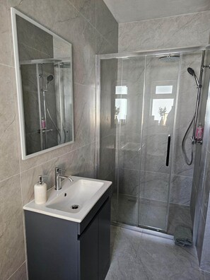 Un baño moderno con un espejo grande, un lavamanos con dispensador de jabón, una ducha con cabezal de ducha de mano y un box de ducha con paredes de vidrio.