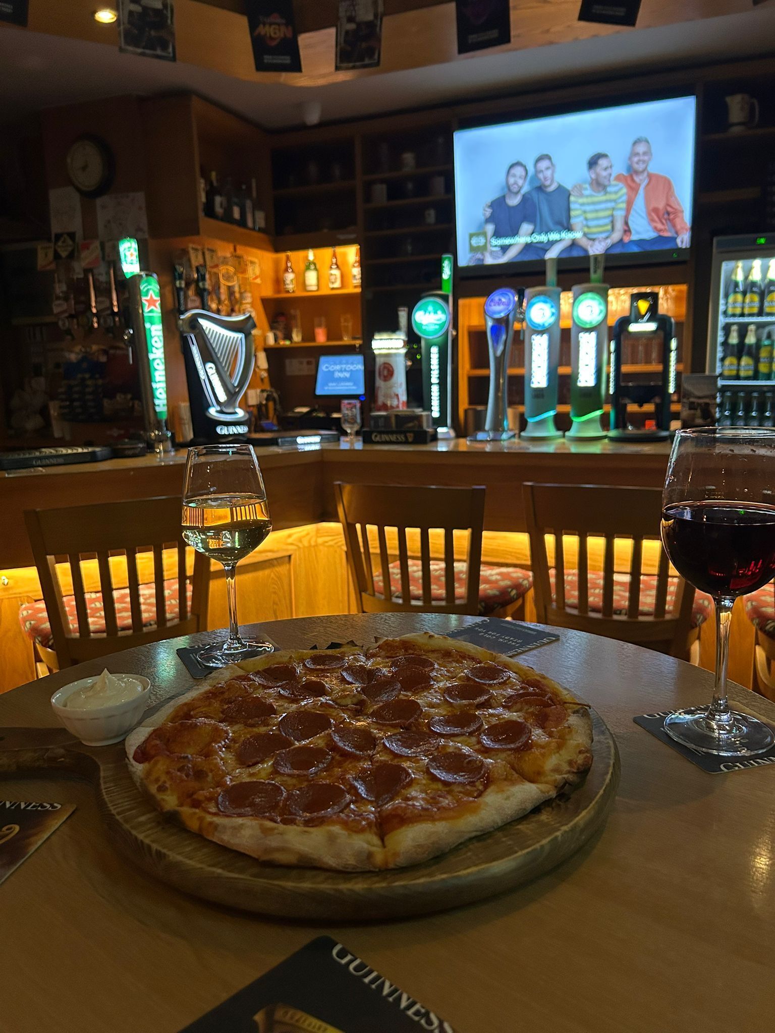 Baaritiskialue, jossa pöydällä on pizza, kaksi lasillista viiniä ja taustalla näkyy televisioruutu.