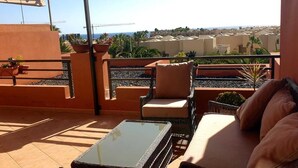 A terrace with a glass table, wicker chairs, and a sofa. There are potted plants and a view of buildings and the sea.