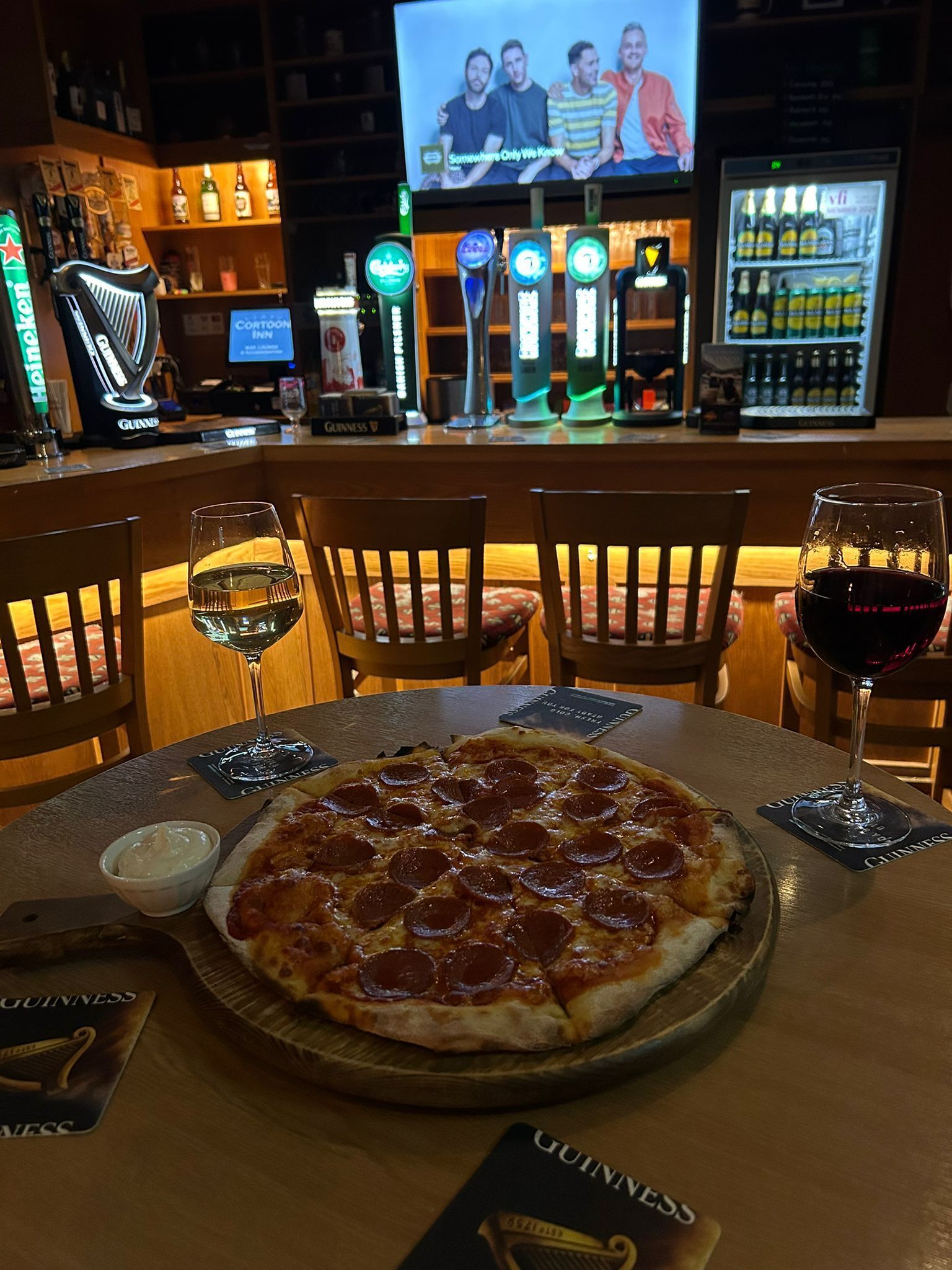 A bar area with a pizza on a table, two glasses of wine, and a television screen in the background.