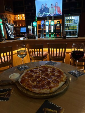 A bar area with a pizza on a table, two glasses of wine, and a television screen in the background. - Cortoon Inn, Cosy Private Twin Room 3, Shared Facilities (Tuam)
