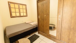 Una habitación pequeña con cama individual, un armario de madera, una ventana con persianas y una mesita.