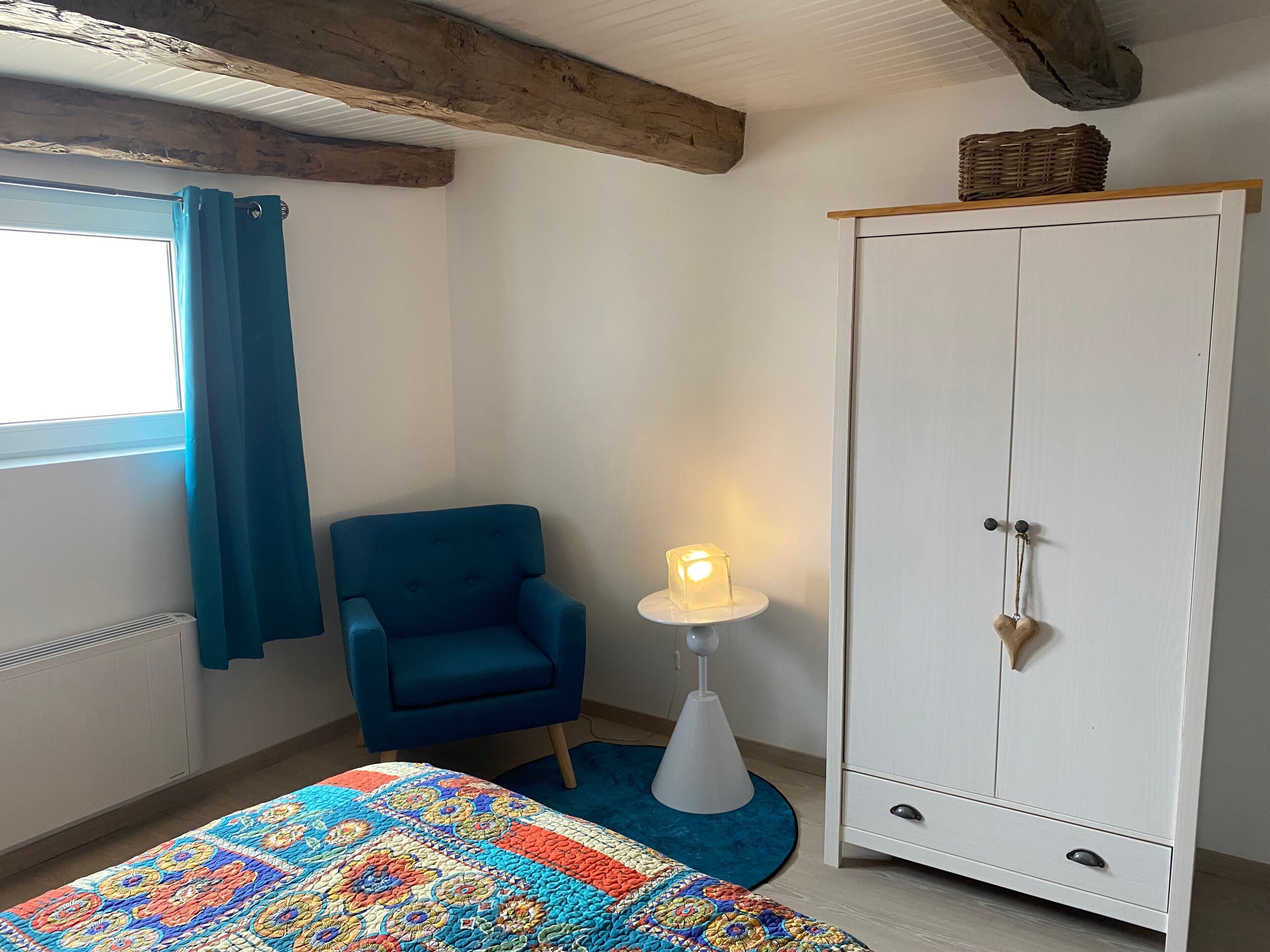 Ein Zimmer mit blauem Sessel, einem weißen Kleiderschrank, einer farbenfrohen Bettdecke und einer Lampe auf einem weißen Beistelltisch.