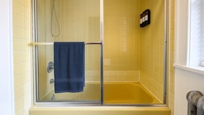 Une douche fermée par des parois en verre, avec des carreaux jaunes, une serviette bleue et une petite étagère sur laquelle sont posées des bouteilles.