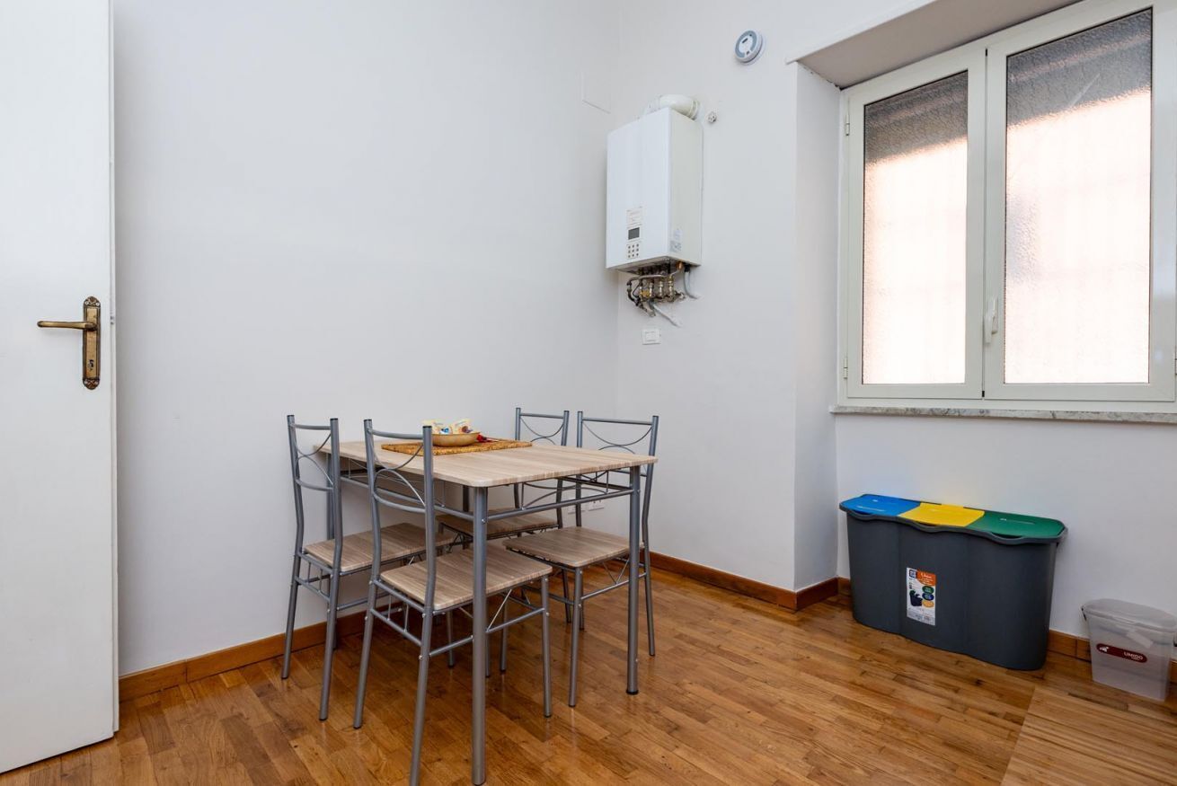 A room with a dining table, chairs, a window, a wall-mounted heater, and a trash bin.