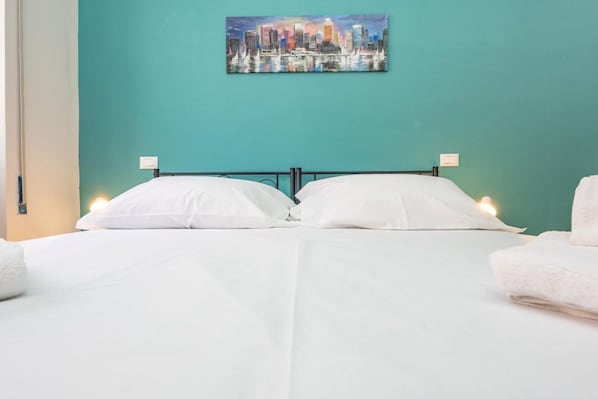 Un lit double avec du linge de lit blanc, un mur de couleur turquoise et, au-dessus, un tableau représentant un paysage urbain.