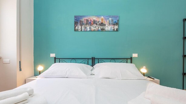 Una cama con sábanas blancas, dos lámparas de noche y una pared color turquesa con un cuadro de paisaje urbano.