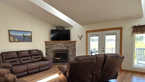 Ein Wohnzimmer mit Kamin aus Stein, Ledersofas, einem Flachbildfernseher und Blick durch eine Glastür.