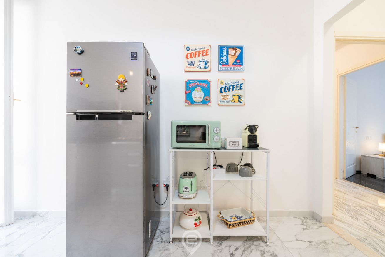 A compact kitchen with a stainless steel refrigerator, a microwave, and a small shelf unit with kitchenware.