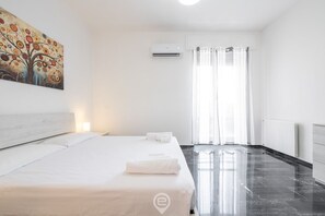 A hotel room with a bed, a painting on the wall, an air conditioning unit, and a view of the outside through a window with curtains.