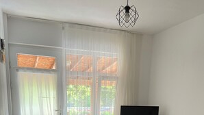 Een woonkamer met een zwart-wit geruit vloerkleed, twee bloemensofa’s, een tv op een standaard, een radiator en een raam met transparante gordijnen.