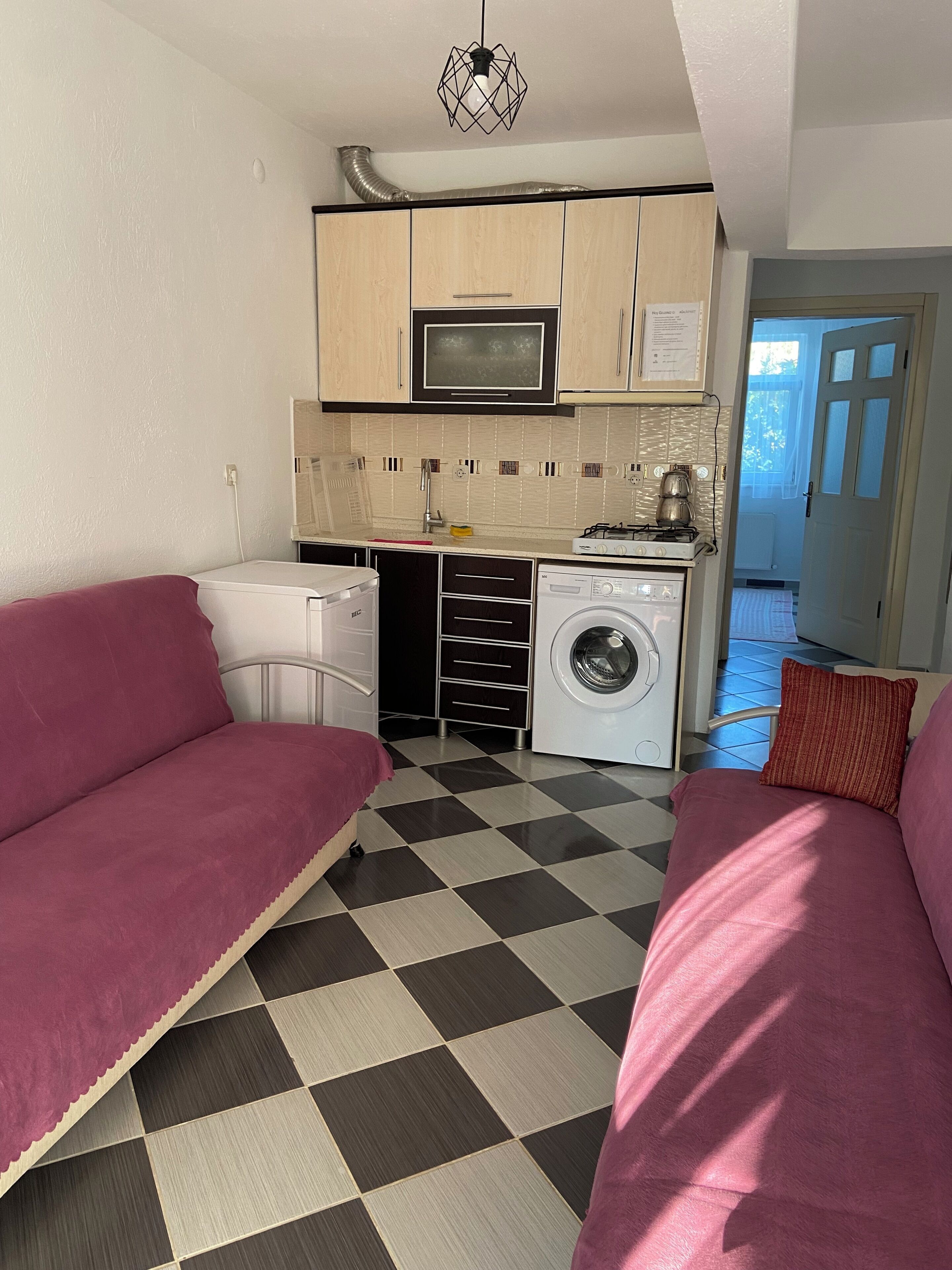Cocina funcional con microondas, estufa y lavadora. Hay un sofá y una cama sobre un piso de cuadros.