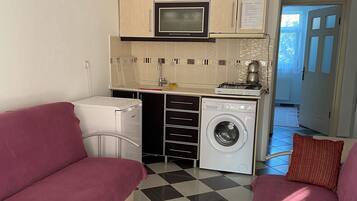 Cocina funcional con microondas, estufa y lavadora. Hay un sofá y una cama sobre un piso de cuadros.