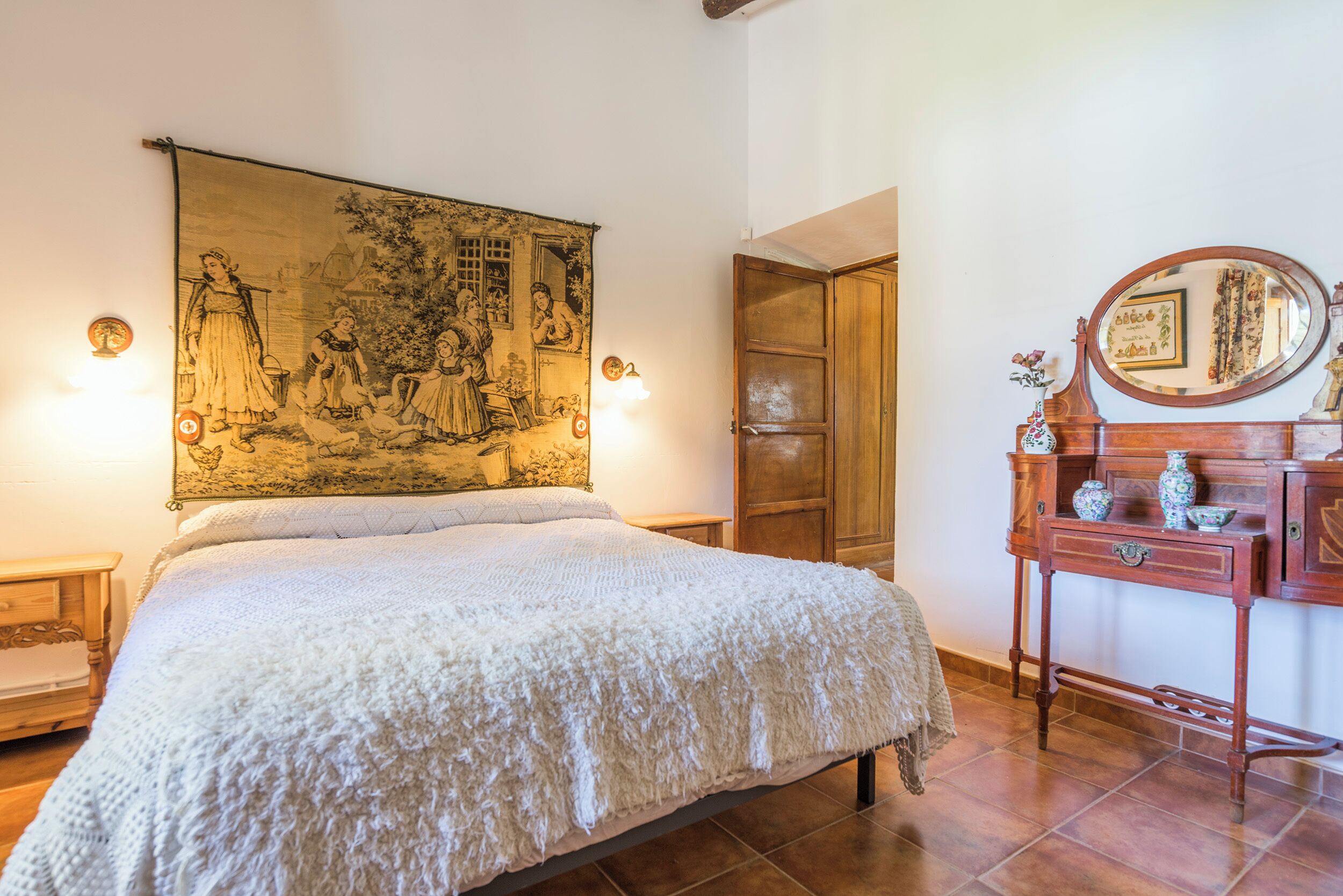 Une chambre à coucher avec une grande tapisserie, une commode en bois avec un miroir et un lit recouvert d’une couverture blanche.