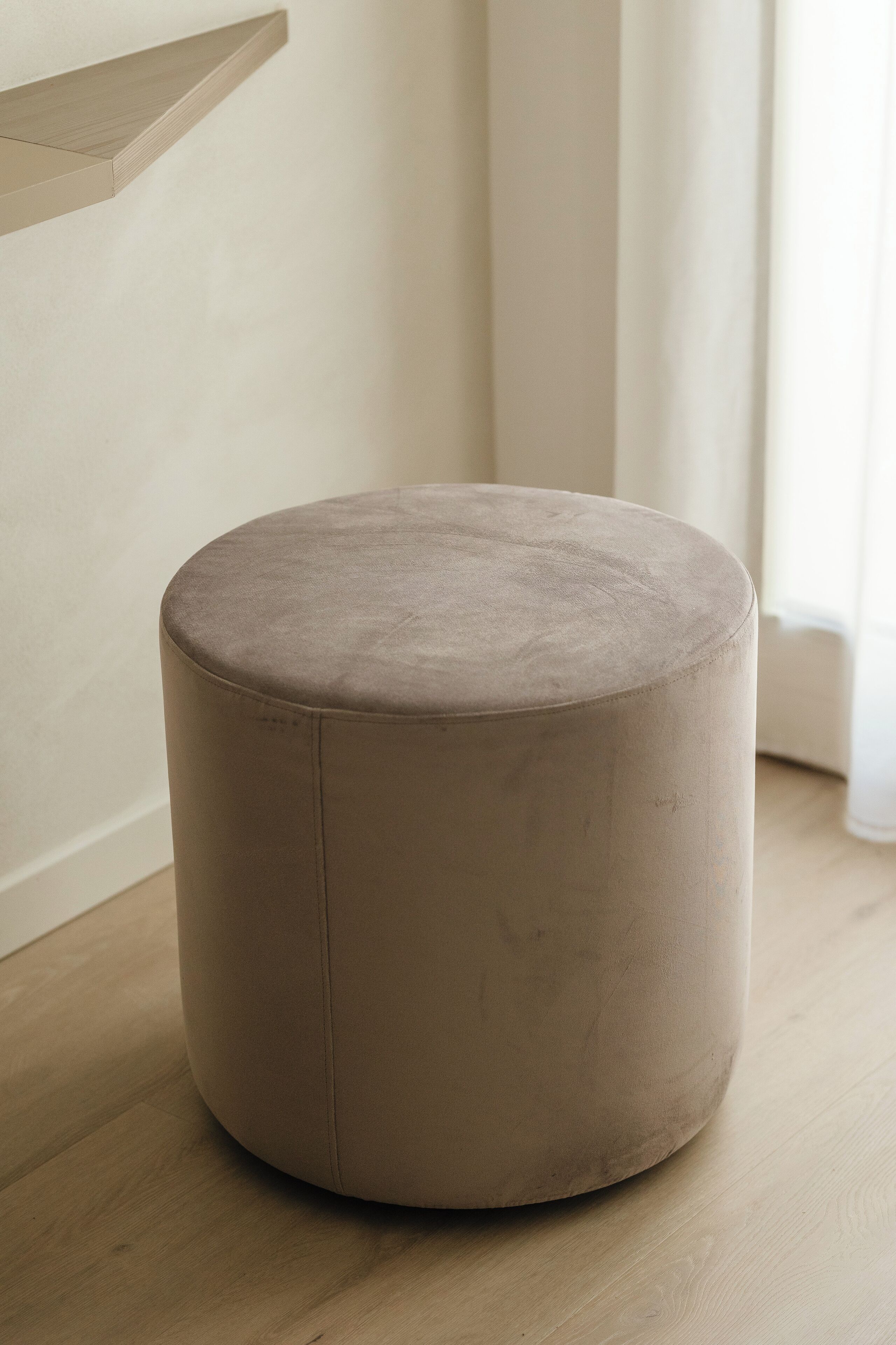 Een rond, beige poef in een kamer met een houten vloer en een witte muur.
